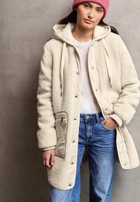 Abrigo largo beige forrado de forro polar, con capucha y broches; presenta un bolsillo de tela beige con cremallera y puños acanalados. Llevado sobre unos vaqueros claros.