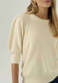 Femme portant un pull en tricot couleur crème avec des manches mi-longues bouffantes et des poignets côtelés, assorti de boucles d'oreilles dorées en forme de créole.