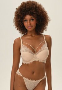 Beżowy stanik koronkowy z falbaniastymi brzegami i ozdobnymi ramiączkami. Pasujące majtki bikini mają podobny wzór koronkowy, podkreślony delikatnymi detalami.