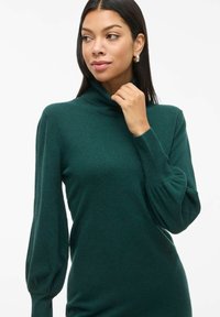 Suéter verde de cuello alto hecho de una tela suave, con mangas abullonadas largas y detalles de ribete en el dobladillo para añadir textura.
