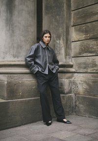 Grau lederne Jacke mit einem strukturierten Kragen, kombiniert mit schwarzen, weit geschnittenen Hosen und spitzen schwarzen Ballerinas, vor einer strukturierten Steinwand.