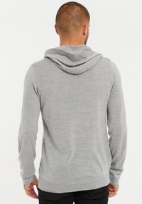 Man met kort haar in een lichtgrijze hoodie en zwarte broek, gezien vanuit de rug tegen een witte achtergrond.