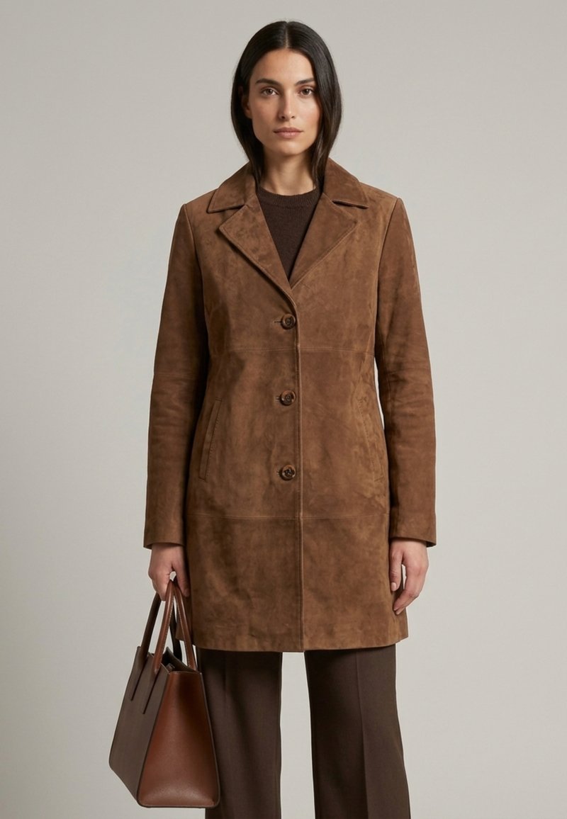 Femme debout, portant un manteau en daim marron boutonné, pantalon foncé, tenant un grand sac à main en cuir marron sur un fond uni.