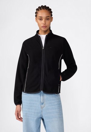 Frau mit geflochtenem Haar trägt eine schwarze Fleecejacke mit Reißverschluss über einem weißen Hemd und hellblauen High-Waist-Jeans vor einem einfarbigen Hintergrund.