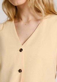 Top di colore crema con design a V, adornato da due bottoni rotondi scuri e un tessuto liscio e leggermente testurizzato.