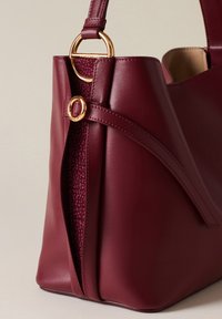 Borsa a spalla in pelle borgogna con fodera in tessuto texturizzato, un anello in metallo dorato e un design distintivo piegato nella parte frontale.