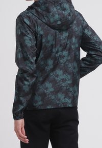 Kurtka z kapturem w abstrakcyjny wzór w ciemnozielonym i czarnym kolorze, wykonana z lekkiego materiału, z elastycznymi mankietami i luźnym krojem.