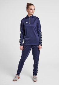 Tuta atletica navy con zip a metà, motivo a chevron contrastante sulle maniche e una texture liscia. Accenti grigi sui pantaloni e sulle scarpe.