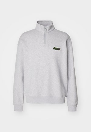 Sudadera de forro polar gris con cuello de media cremallera y puños de canalé, que presenta un logo de cocodrilo bordado en verde en el lado izquierdo del pecho.