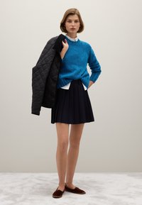 Gebreide blauwe trui, witte blouse met kraag eronder, zwarte doorgestikte jas over één arm gedrapeerd, geplooide marineblauwe rok, bruine instapschoenen.