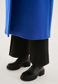 Botines de cuero negro con suelas gruesas, combinados con pantalones sueltos negros recortados bajo un abrigo azul brillante. Textura lisa y herrajes mínimos.