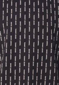 Svart tyg med upprepande vit text som säger "TOMMY JEANS" i ett vertikalt och horisontellt mönster. Slät yta, töjbart material.