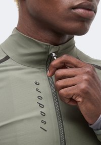 Gros plan sur la main d'une personne tirant la fermeture éclair d'une veste vert olive avec « Isadore » imprimé verticalement.