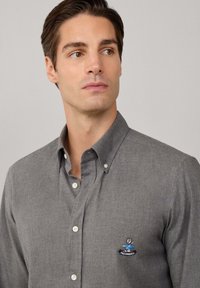 Hombre con cabello castaño que lleva una camisa gris abotonada con un pequeño logo bordado de Façonnable en el pecho, mirando hacia un lado.