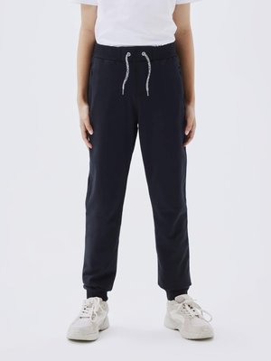 Calças de treino azul marinho com cintura ajustável por cordão, design elegante, bolsos laterais e punhos ajustados; combinadas com ténis de cor clara.