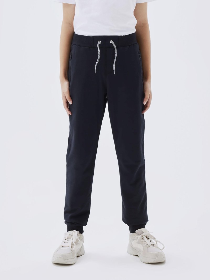 Navyblå joggebukser med snor i midjen, slank design, sidelommer og tettsittende mansjetter; kombinert med lyse sneakers.