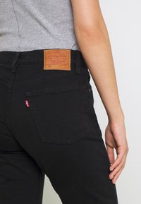 Jeans in denim nero con taglio classico, caratterizzati da una patta in pelle sulla parte posteriore della vita e un piccolo logo rosso sulla tasca posteriore.
