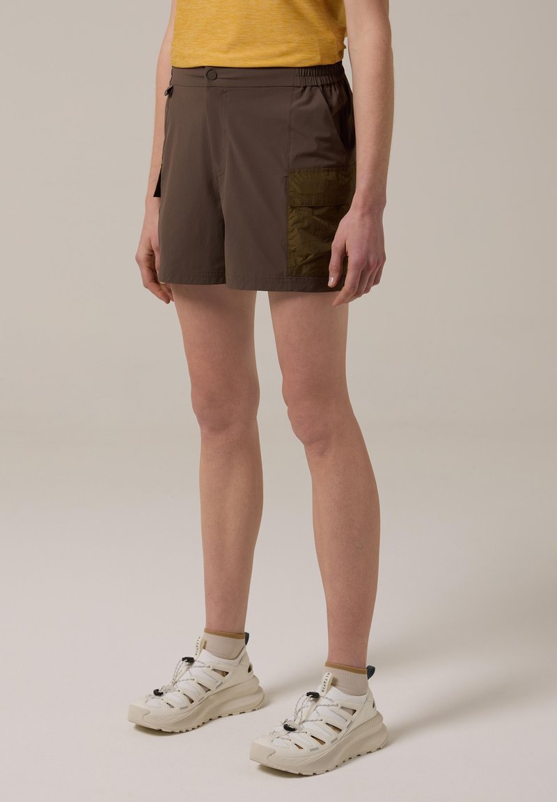 Persona con pantalones cortos marrones con bolsillo lateral, camiseta amarilla, calcetines beige y zapatillas deportivas blancas, de pie sobre un fondo neutro.