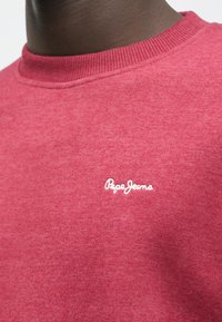 Felpa rossa con scollatura rotonda a costine, realizzata in tessuto testurizzato. Presenta un logo "Pepe Jeans" ricamato sul petto.