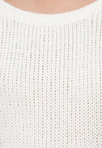 Biały sweter z dzianiny o teksturowanym wzorze z małymi brązowymi plamkami, okrągłym dekoltem i prążkowanymi brzegami. Widoczny miękki materiał.