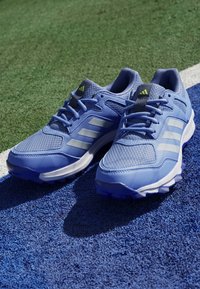 Modré atletické topánky s sieťovaným zvrškom, tromi bielymi pruhmi, gumovou podrážkou a polstrovaným golierom, umiestnené na modrom textúrovanom povrchu.