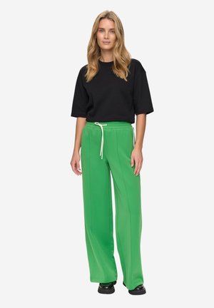 Čierne oversize tričko s krátkymi rukávmi v kombinácii s vysokými zelenými teplákmi. Nohavice majú šnúrku v páse a široký strih nohavíc.