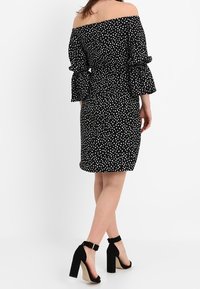 Svart off-shoulder klänning med vita prickar, utsvängda ärmar och en figurnära silhuett, matchad med svarta högklackade sandaler.