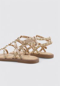 Sandalias beige con tachuelas y acentos en dorado, con tiras entrecruzadas y suela plana. Fabricadas en material sintético con una textura suave.