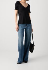 T-shirt nero a maniche corte con scollo in pizzo, abbinato a jeans a gamba larga di un azzurro chiaro. Il modello tiene in mano una borsa nera.