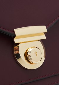 Dettaglio di una borsa in pelle bordeaux con un lucchetto rotondo e lucido color oro con una serratura, accentuata da una chiusura liscia in oro e cuciture coordinate.