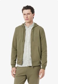 Felpa olive green con zip in tessuto morbido, caratterizzata da una tasca a marsupio e cappuccio con cordino. Indossata sopra una camicia a strisce chiara.