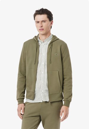 Felpa olive green con zip in tessuto morbido, caratterizzata da una tasca a marsupio e cappuccio con cordino. Indossata sopra una camicia a strisce chiara.