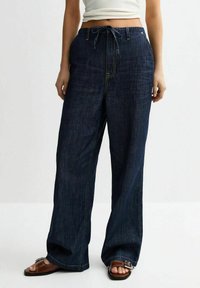 Pantalons en denim sombre à jambes larges avec une taille à cordon de serrage, présentant de subtils accents décolorés et des coutures doubles pour une durabilité accrue.