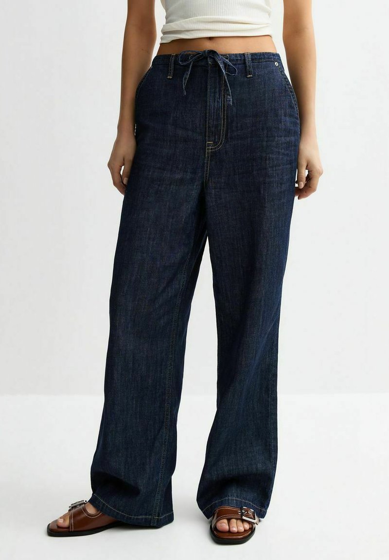 Pantalons en denim sombre à jambes larges avec une taille à cordon de serrage, présentant de subtils accents décolorés et des coutures doubles pour une durabilité accrue.
