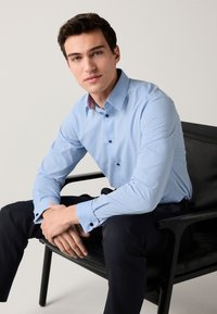 Šviesiai mėlyna marškiniai su tekstūruotu raštu, kontrastingu raštuotu apykakle ir juodais rankogalių mygtukais. Modelis sėdi, dėvi tamsias kelnes.