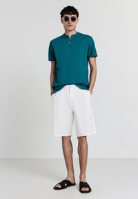 Chemise henley à manches courtes teal avec un bouton, associée à un short blanc au genou et des sandales noir à enfiler. Tissu lisse et coupe décontractée.