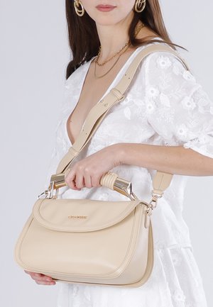 Vrouw in witte jurk met bloemenprint, die een beige lederen handtas vasthoudt met een gouden handvat en een verstelbare schouderriem.