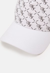 Hvid kasket med et gentagende sort dekorativt mønster med logoelementer og krydsende linjer. Blødt stof og struktureret skygge.