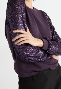 Top de manga larga morado con mangas adornadas con lentejuelas, con un cuerpo de tela suave y puños de canalé. Combinado con pantalones negros.