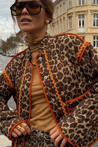 Jasje met luipaardprint, gewatteerd en met oranje accenten, met drukknopen aan de voorkant, en bijpassende broek. Beigekleurige coltrui eronder; gouden ringen aan de hand.
