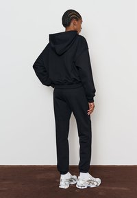 Hanorac negru și pantaloni sport dintr-un material moale, cu croială relaxată. Are glugă și manșete elastice, asortate cu adidași albi și argintii.