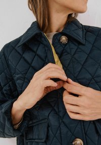 Marineblå quilted jakke med en gul foring, der har en bred krave, fremtrædende knapper og sidelommer. Glat tekstur med et diamantmønster.