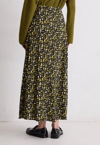 Vrouw in een geklede lange rok met zwarte, olijfgroene en witte blokken, olijfgroene trui en zwarte loafers, staande op een witte vloer.