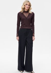 Brązowa bluzka z długim rękawem z koronkowymi akcentami przy dekolcie i mankietach, zestawiona z czarnymi spodniami o szerokich nogawkach. Spodnie mają gładką, płynną fakturę.