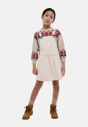 Fille debout, mains relâchées, portant une robe salopette en velours côtelé beige, un pull fleuri, des chaussettes beiges et des bottines marron à lacets.