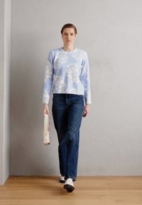Lyseblå genser med blomsterdesign, parret med mørkeblå jeans. Modellen holder en hvit håndveske og har på seg hvite sko. Bakgrunn med tregulv.