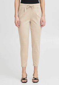 Beige, taps toelopende broeken met een trekkoord in de taille, voorplooien, een gladde textuur en een enkel-lange pasvorm. Gepaarde met zwarte sandalen.