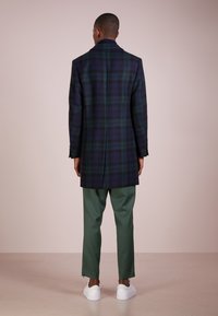 Lungo cappotto scozzese in verde scuro e blu navy, con una vestibilità sagomata, collo a rever e due spacchi posteriori, abbinato a pantaloni verdi e scarpe bianche.