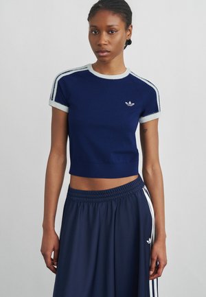 Frau trägt navyblaues, kurz geschnittenes Adidas T-Shirt und passende Trainingshose mit weißen Streifen und steht vor einem schlichten grauen Hintergrund.