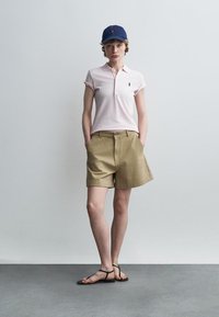 Vrouw met een marineblauwe pet, lichtroze poloshirt, kaki korte broek en zwarte sandalen met bandjes, staand met handen in de zakken.